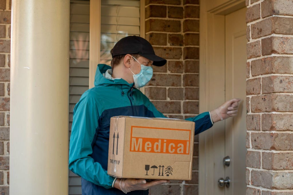 Contactless delivery during COVID-19 pandemic lockdown concept. Courier wearing mask and gloves holds a parcel with medical equipment and knocks on the door.